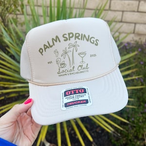 May include: A beige trucker hat with a green embroidered logo that says "Palm Springs Social Club est 2025 Naomi's Bachelorette". The logo also features a flamingo, palm trees, and a cocktail glass. The hat has a black and white Otto label that says "World Famous Foam Front OSFM Trucker Durable Snap Closure".