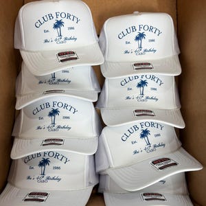 May include: A stack of white trucker hats with a blue palm tree graphic and the text "CLUB FORTY Est. 1986 Bri's 40th Birthday CABO". The hats have a mesh back and a curved brim.