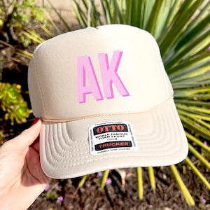 May include: A beige trucker hat with a pink "AK" logo on the front. The hat has a mesh back and a snap closure. The hat is made by OTTO and says "World Famous Foam Front" on the label.