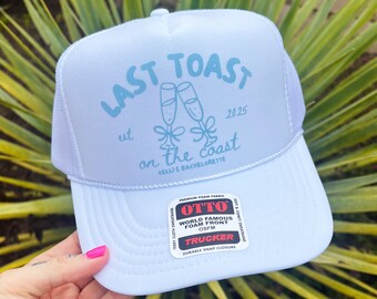 May include: A light blue trucker hat with a white mesh back. The hat features the text "LAST TOAST on the coast 2025" in light blue, with champagne glasses graphic. The hat has a snap closure and a "OTTO" label.
