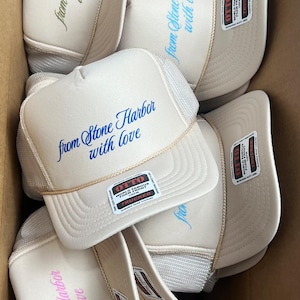 May include: A collection of beige trucker hats with white mesh backs. Each hat features the embroidered text "from Stone Harbor with love" in various colors. The hats have a curved brim and a snap closure.