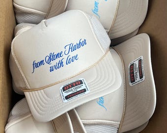 May include: A collection of beige trucker hats with mesh backs and curved brims. Each hat features the embroidered text "from Stone Harbor with love" in various colors. Some hats have an "OTTO" label.