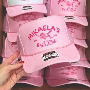 May include: A light pink trucker hat featuring a foam front with dark pink text that reads MIKAELA'S est. 2026 Bach Club CHARLESTON and a graphic of a person in a martini glass. The hat includes an Otto brand sticker on the brim and a snap closure.