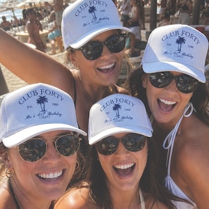 May include: Four smiling people wearing white trucker hats and sunglasses on a sunny beach. The hats read "CLUB FORTY Est. 1986 Bri's 40th Birthday CABO" in blue text. The background shows a beach scene with umbrellas and other people.