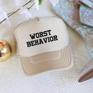 May include: A beige trucker hat with a black mesh back. The front of the hat has the text "WORST BEHAVIOR" in black block letters.
