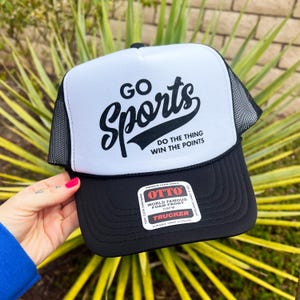 May include: A white and black trucker hat with a black mesh back. The front of the hat has the text "GO Sports" in a black cursive font. Below the text is the phrase "DO THE THING WIN THE POINTS". The hat has a black snap closure and a label that reads "OTTO WORLD FAMOUS FOAM FRONT OSFM TRUCKER DURABLE SNAP CLOSURE".