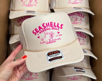 Op de afbeelding: Een stapel beige truckerpetten met een mesh achterkant en een gebogen klep. Elke pet heeft de roze tekst "SEASHELLS & WEDDING BELLS", samen met andere tekst en afbeeldingen. De petten zijn gestapeld in een kartonnen doos.