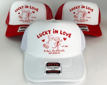 Puede incluir: Gorra de camionero blanca y roja con el texto "Lucky in Love" en rojo. La gorra también tiene el texto "Kaley's Bachelorette, Las Vegas, NV" en rojo. La gorra tiene una parte trasera de malla blanca y una parte delantera roja.
