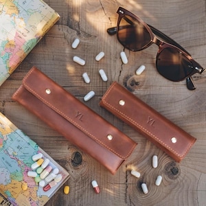 May include: Two brown leather cases with the initials "YL" embossed on the front, along with a pair of sunglasses and scattered pills on a wooden surface. The cases have a flap closure with gold-colored studs. A map is visible in the background.