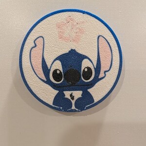 Może przedstawiać: Okrągły niebiesko-biały magnes z nadrukowaną w 3D grafiką postaci Stitcha. Projekt zawiera niebieską obwódkę, białe tło, jasnoróżowy kwiat hibiskusa nad postacią oraz Stitcha z dużymi uszami i oczami.