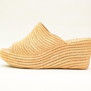 May include: A woven raffia wedge sandal in a natural beige color. The open-toe sandal has a curved platform sole and a woven upper that covers the top of the foot. The heel measures approximately 3 inches.