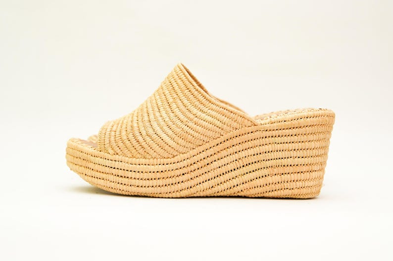 May include: A woven raffia wedge sandal in a natural beige color. The open-toe sandal has a curved platform sole and a woven upper that covers the top of the foot. The heel measures approximately 3 inches.