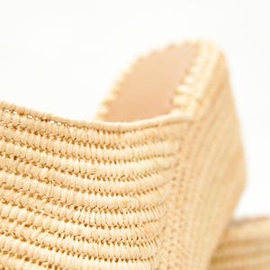 May include: Close-up of a woven raffia shoe. The shoe is a light beige color with a textured surface. The interior of the shoe is a lighter beige color. The shoe is set against a white background.