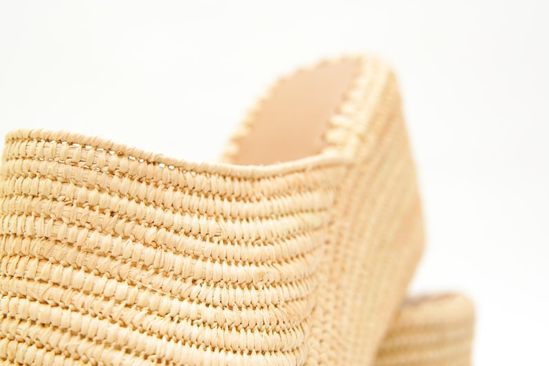 May include: Close-up of a woven raffia shoe. The shoe is a light beige color with a textured surface. The interior of the shoe is a lighter beige color. The shoe is set against a white background.