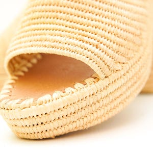 May include: Close-up of a pair of woven raffia sandals. The open-toe design reveals a leather insole. The sandals have a platform sole and a woven texture, showcasing a natural, beige color. The focus is on the intricate weaving.