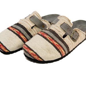 May include: A pair of beige clogs with a striped pattern and buckle fastening. The clogs feature a woven fabric upper with red, black, and grey stripes. The soles are black with a cork footbed and a grey strap.