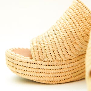 May include: Close-up of a woven raffia platform sandal. The sandal features a thick, textured sole and a woven upper. The natural beige color of the raffia contrasts with the light pink interior. The design suggests a summer or beach theme.