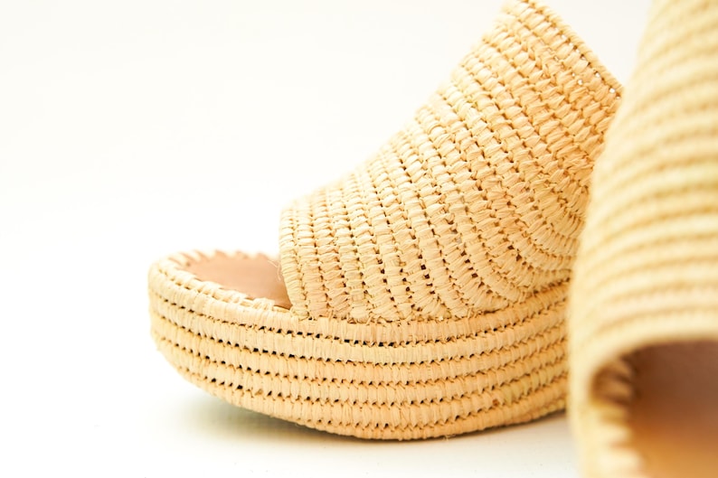 May include: Close-up of a woven raffia platform sandal. The sandal features a thick, textured sole and a woven upper. The natural beige color of the raffia contrasts with the light pink interior. The design suggests a summer or beach theme.