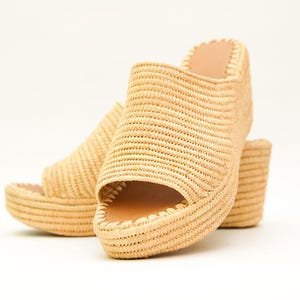 May include: A pair of woven raffia platform sandals. The open-toe shoes have a thick sole and a small heel. The natural beige color of the raffia contrasts with the light brown interior of the shoe.