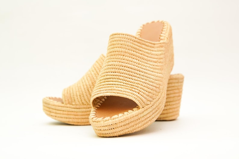 May include: A pair of woven raffia platform sandals. The open-toe shoes have a thick sole and a small heel. The natural beige color of the raffia contrasts with the light brown interior of the shoe.