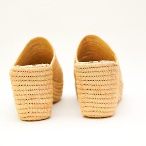 May include: Two pairs of woven raffia slip-on sandals. The open-toe shoes have a natural beige color and a textured, basket-weave design. The shoes are set against a white background.