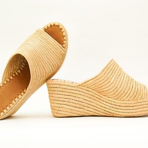 May include: Two woven raffia sandals. One is a flat slide with a light brown leather insole and a stitched edge. The other is a wedge sandal with an open toe and a woven upper. Both are beige.
