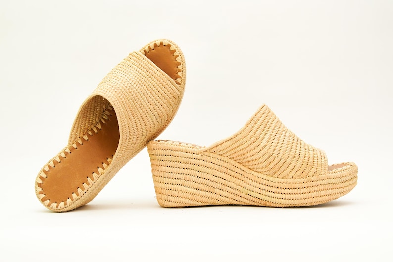 May include: Two woven raffia sandals. One is a flat slide with a light brown leather insole and a stitched edge. The other is a wedge sandal with an open toe and a woven upper. Both are beige.