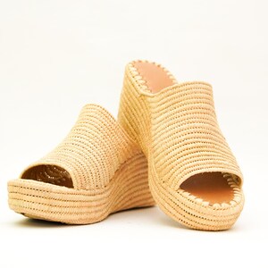 May include: A pair of beige woven wedge sandals with open toes. The sandals have a platform sole and a woven upper. The interior is a light brown color. The shoes are set against a white background.