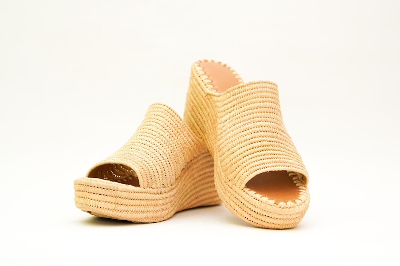 May include: A pair of beige woven wedge sandals with open toes. The sandals have a platform sole and a woven upper. The interior is a light brown color. The shoes are set against a white background.