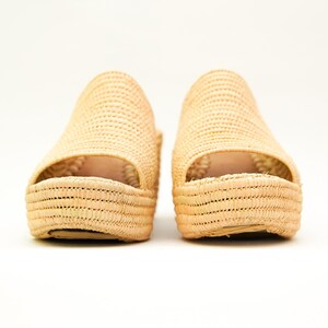 May include: A pair of natural woven raffia platform sandals. The open-toe shoes have a woven texture and a thick sole. The sandals are beige and sit on a white background.