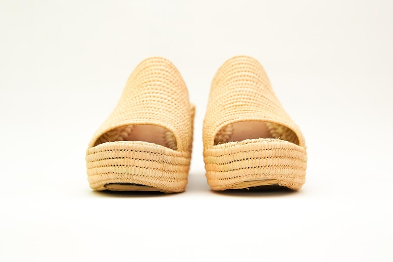 May include: A pair of natural woven raffia platform sandals. The open-toe shoes have a woven texture and a thick sole. The sandals are beige and sit on a white background.