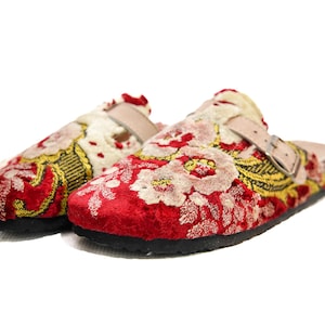Könnte beinhalten: Ein Paar rot bestickte Clogs mit Blumenmustern in Weiß und Gold. Die Clogs haben eine schwarze Sohle, einen beigefarbenen Riemen mit Schnalle und ein Korkfußbett. Die Schuhe sind vor weißem Hintergrund.