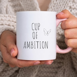 May include: A white ceramic mug with a light pink handle. The front of the mug features the black text "CUP OF AMBITION" written in a simple sans-serif font, accompanied by a small, minimalist black line drawing of a butterfly to the right.
