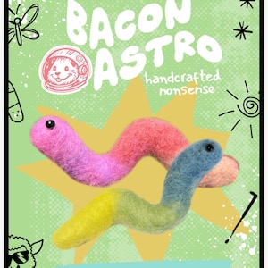 May include: A needle felting kit package featuring a green background with a yellow star and two felted worm figures, one pink and one blue and yellow. The text reads Bacon Astro, Stab Through The Scroll, and Needle Felting Kit For The Craft-Curious.