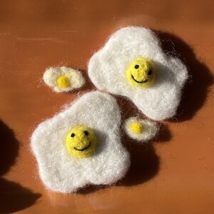 May include: Two medium-sized felted egg shapes with white whites and yellow yolks, each featuring a black embroidered smiley face. Two smaller, plain felted egg shapes without faces are positioned nearby, all displaying a soft, fuzzy texture.