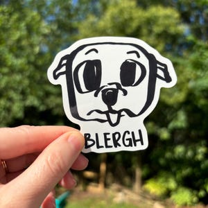 Op de afbeelding: Een witte sticker met een cartoon hondengezicht met grote ogen, een kleine neus en een tong. Het woord "BLERGH" is in zwart onder het gezicht van de hond gedrukt. De sticker wordt tegen een vage groene achtergrond gehouden.