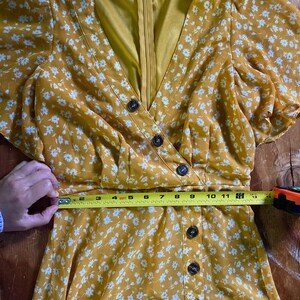 May include: A yellow floral dress with white flowers and a button-up front. The dress has short sleeves and a V-neckline. A measuring tape is placed around the waist of the dress, showing a measurement of 30 inches.