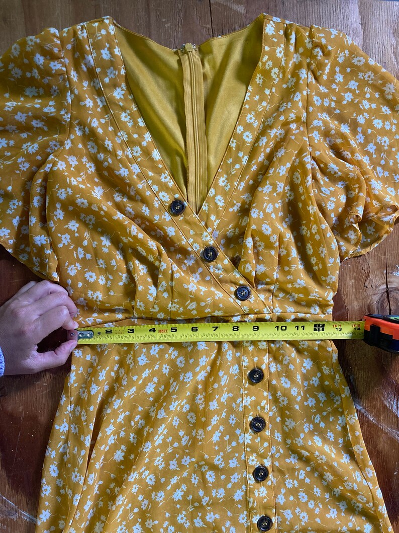 May include: A yellow floral dress with white flowers and a button-up front. The dress has short sleeves and a V-neckline. A measuring tape is placed around the waist of the dress, showing a measurement of 30 inches.