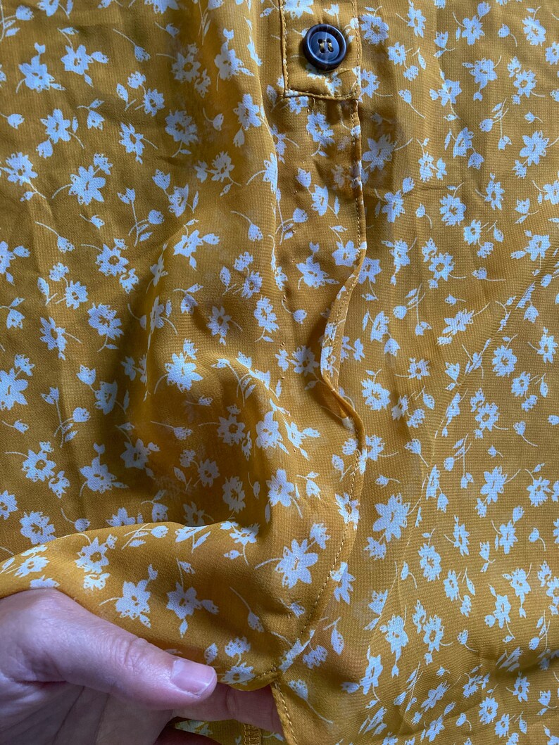 May include: A yellow sheer blouse with a white floral pattern. The blouse has a button closure at the neck.