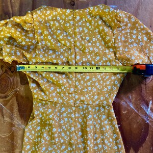 May include: A yellow dress with a white floral pattern. The dress has a V-neckline and short sleeves. The dress is measured across the back with a tape measure, showing a measurement of 18 inches.