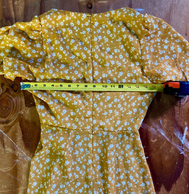 May include: A yellow dress with a white floral pattern. The dress has a V-neckline and short sleeves. The dress is measured across the back with a tape measure, showing a measurement of 18 inches.
