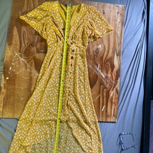 May include: A yellow maxi dress with a white floral print and a button-up front. The dress has short sleeves and a V-neckline. The dress is measured with a tape measure, showing a length of 48 inches.
