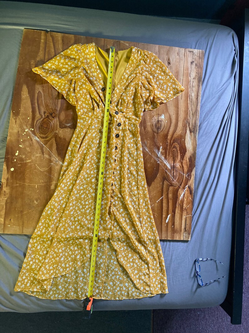 May include: A yellow maxi dress with a white floral print and a button-up front. The dress has short sleeves and a V-neckline. The dress is measured with a tape measure, showing a length of 48 inches.