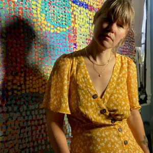May include: A woman wearing a yellow floral dress with white buttons and a silver necklace stands in front of a colorful abstract painting. The painting is made up of small, colorful dots.