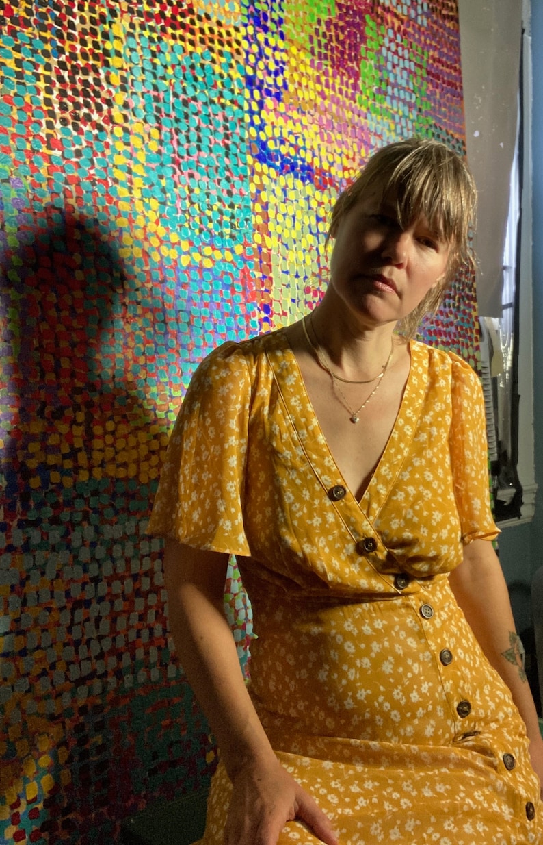May include: A woman wearing a yellow floral dress with white buttons and a silver necklace stands in front of a colorful abstract painting. The painting is made up of small, colorful dots.