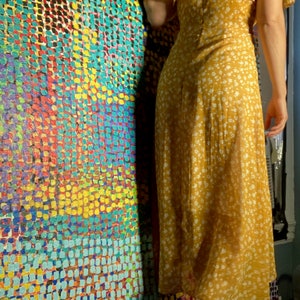 May include: A woman wearing a yellow floral maxi dress with a white background. The dress has a V-neckline and short sleeves. She is standing in front of a wall with a colorful abstract pattern.