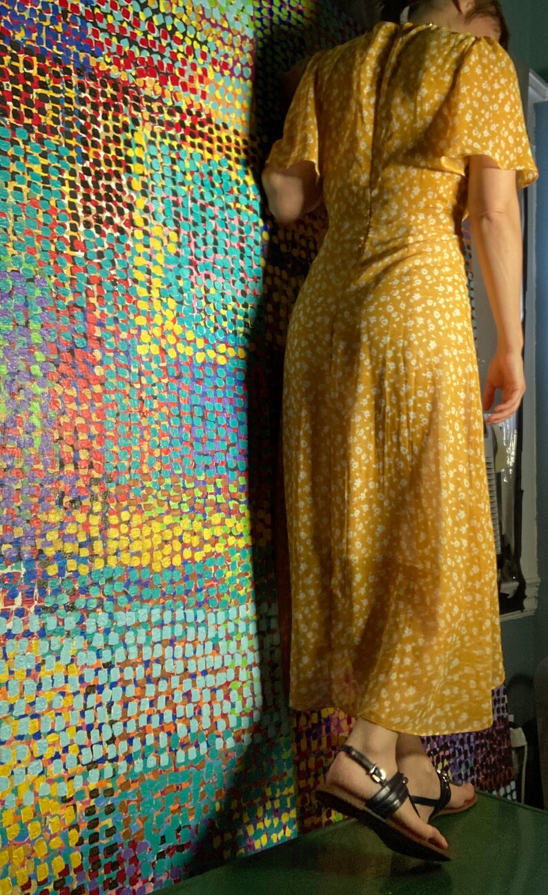 May include: A woman wearing a yellow floral maxi dress with a white background. The dress has a V-neckline and short sleeves. She is standing in front of a wall with a colorful abstract pattern.