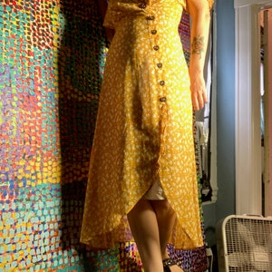 May include: A yellow floral maxi dress with a wrap style and button closure. The dress has a high neckline and short sleeves. The model is wearing the dress with black sandals.