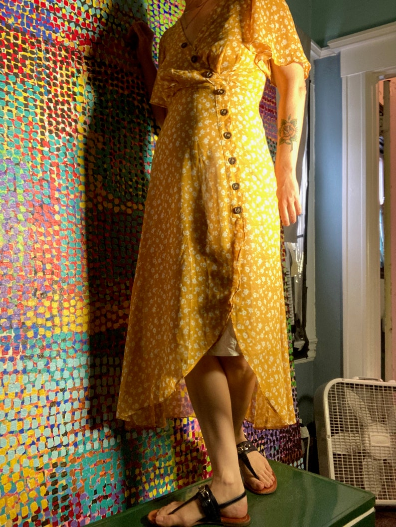 May include: A yellow floral maxi dress with a wrap style and button closure. The dress has a high neckline and short sleeves. The model is wearing the dress with black sandals.