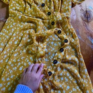 May include: A yellow floral wrap dress with white flowers and a row of buttons down the front.